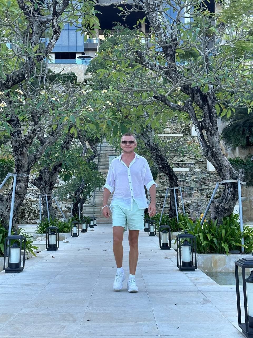 Stylish man in white shirt and green shorts walks tropical path with lanterns.