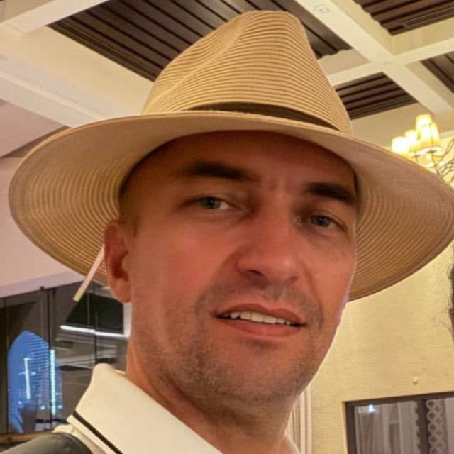 A man with stubble wearing a tan wide-brimmed hat and white polo shirt, smiling.